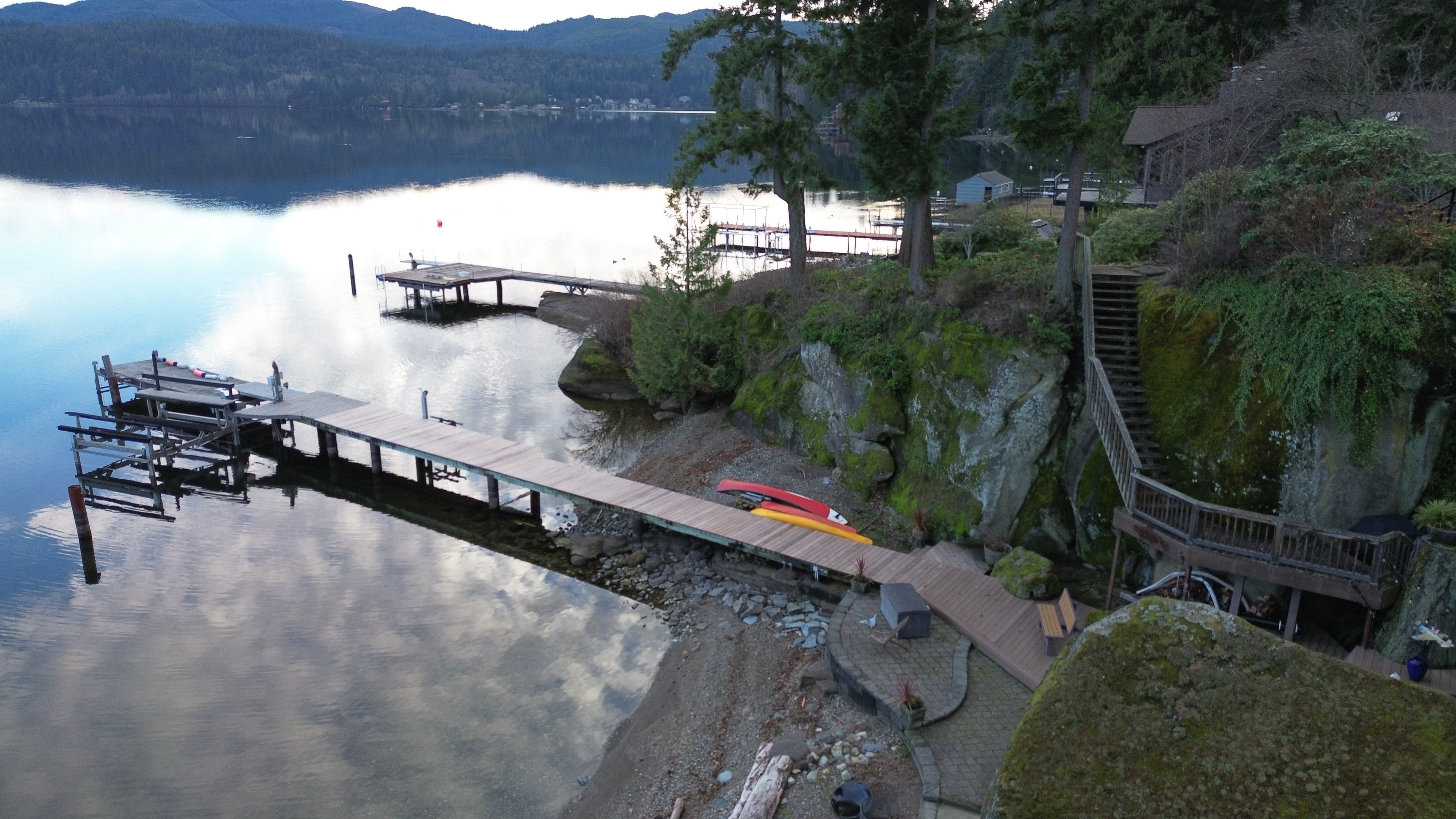  clouds reflected in lake surround Ipe dock in Bellingham, WA. evergreen trees grow along the cliff side stairway.