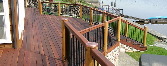 Big Lake, Skagit County Brazilian Redwood Deck with custom railing. the decking is fastened with stainless steel screws wich were pluged and sanded after install. The railing is iron picket panels attached to round cedar posts with copper poct caps, the top rail is Brazilian Redwood to match the decking.